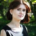 A smiling woman outside, as the sun shines, trees are seen in the background.